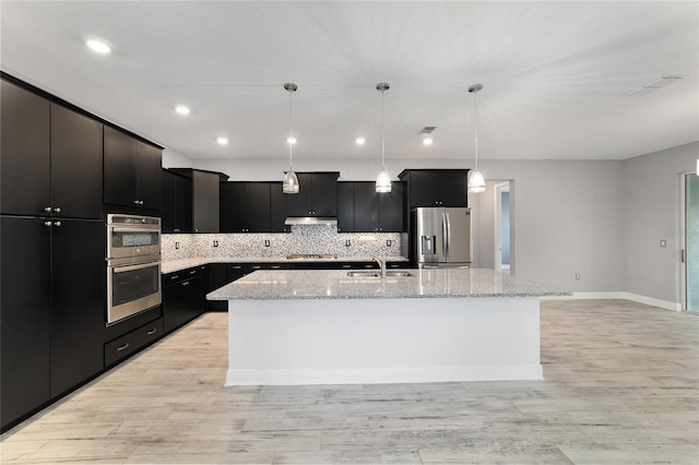 kitchen featuring an island with sink, hanging light fixtures, stainless steel appliances, dark cabinetry, and a sink