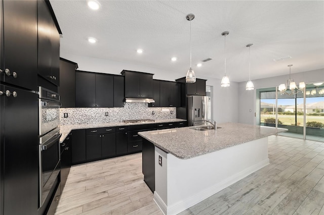 kitchen featuring pendant lighting, appliances with stainless steel finishes, a kitchen island with sink, dark cabinets, and under cabinet range hood