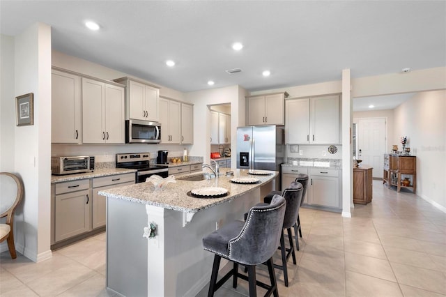 kitchen featuring an island with sink, a breakfast bar, stainless steel appliances, gray cabinetry, and a sink