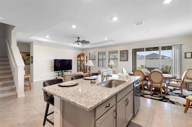 kitchen with ceiling fan, a sink, dishwasher, a raised ceiling, and a center island with sink