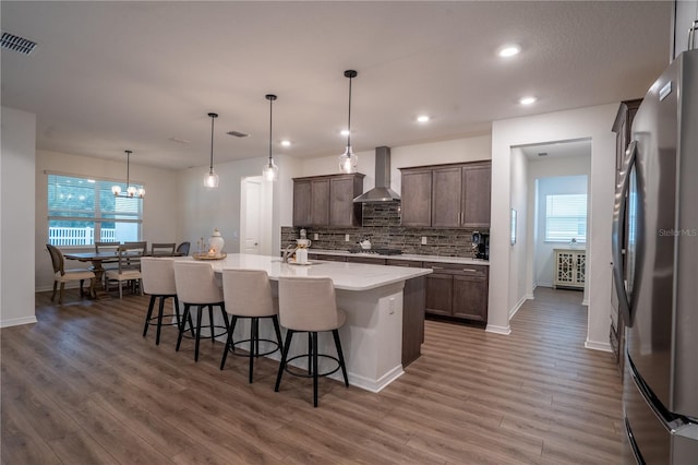 kitchen featuring pendant lighting, appliances with stainless steel finishes, dark brown cabinets, an island with sink, and wall chimney exhaust hood