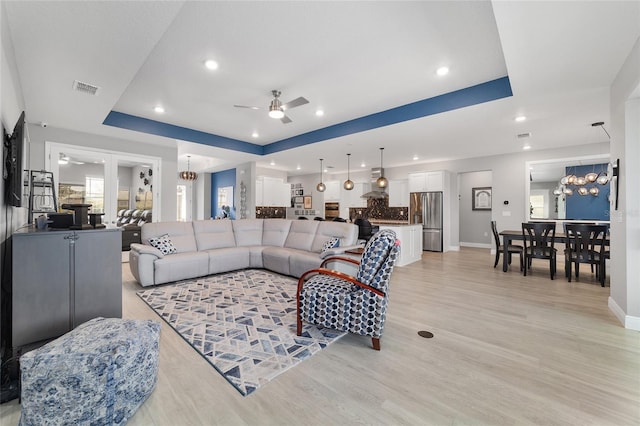 living room featuring a raised ceiling, ceiling fan with notable chandelier, and light hardwood / wood-style flooring