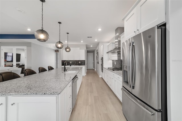 kitchen featuring a spacious island, sink, wall chimney range hood, stainless steel appliances, and white cabinets