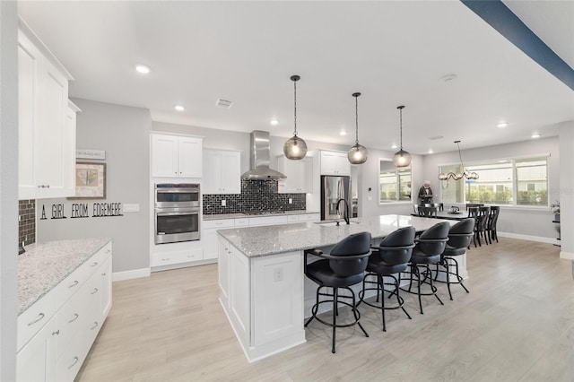 kitchen featuring appliances with stainless steel finishes, white cabinets, a large island with sink, light stone counters, and wall chimney exhaust hood