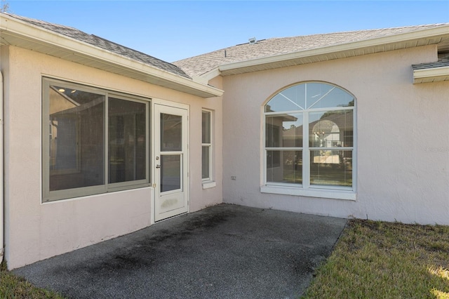 entrance to property with a shingled roof, a patio area, and stucco siding