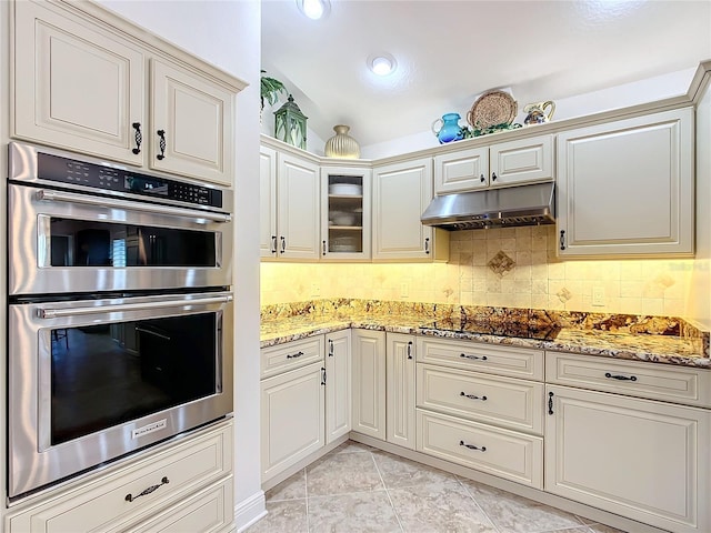 kitchen featuring black electric stovetop, glass insert cabinets, stainless steel double oven, light stone countertops, and under cabinet range hood