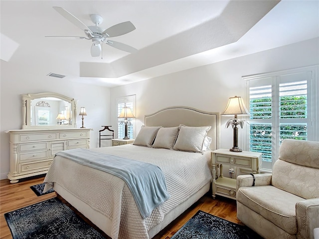 bedroom with a ceiling fan, a tray ceiling, wood finished floors, and visible vents