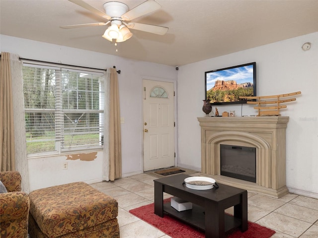 living room featuring baseboards, light tile patterned flooring, a ceiling fan, and a glass covered fireplace