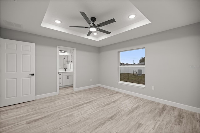 unfurnished bedroom with light wood finished floors, recessed lighting, a raised ceiling, ensuite bath, and baseboards