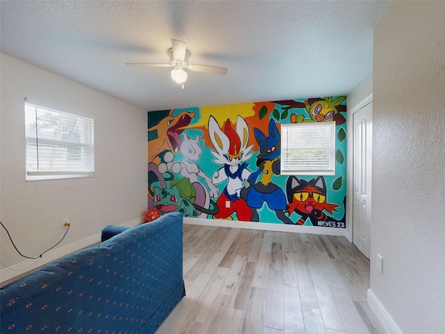 recreation room featuring a textured ceiling, an accent wall, a ceiling fan, baseboards, and light wood-type flooring