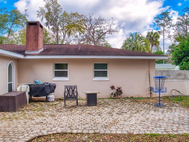 rear view of property with a shingled roof, a patio, a chimney, fence, and stucco siding