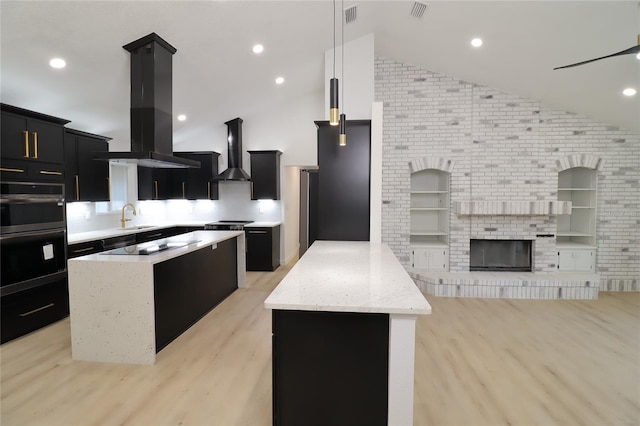 kitchen featuring a large fireplace, wall chimney range hood, dark cabinetry, a center island, and island exhaust hood