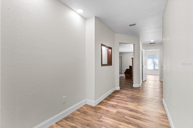 hall featuring a textured ceiling, a textured wall, visible vents, baseboards, and light wood-style floors