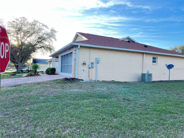 view of side of home with roof with shingles, central AC, a lawn, and stucco siding