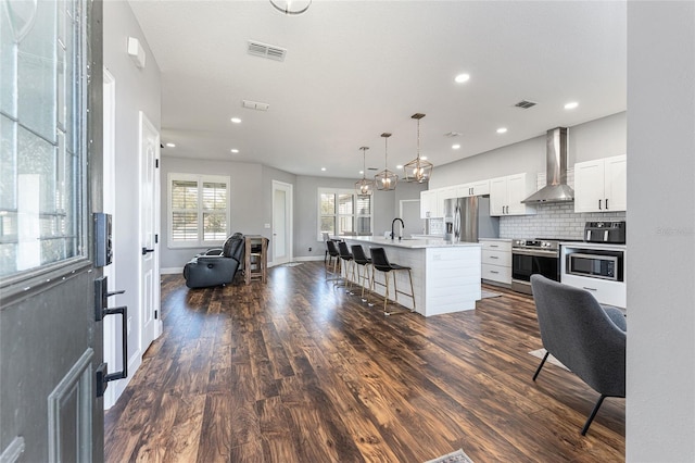 kitchen featuring an island with sink, appliances with stainless steel finishes, light countertops, wall chimney range hood, and pendant lighting