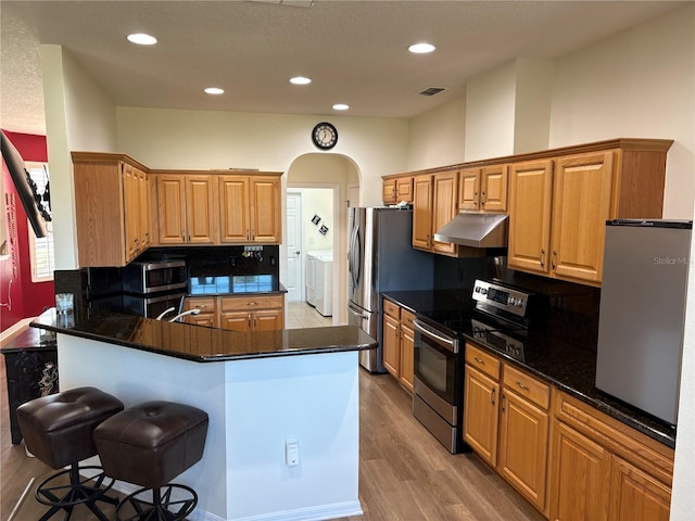 kitchen featuring visible vents, appliances with stainless steel finishes, washing machine and clothes dryer, under cabinet range hood, and a kitchen bar