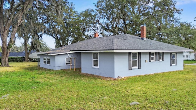 back of house with stucco siding, roof with shingles, a chimney, and a yard