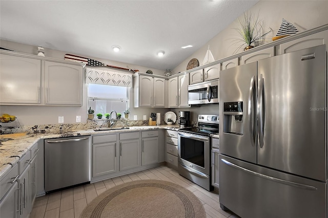 kitchen with lofted ceiling, light stone counters, gray cabinetry, a sink, and appliances with stainless steel finishes