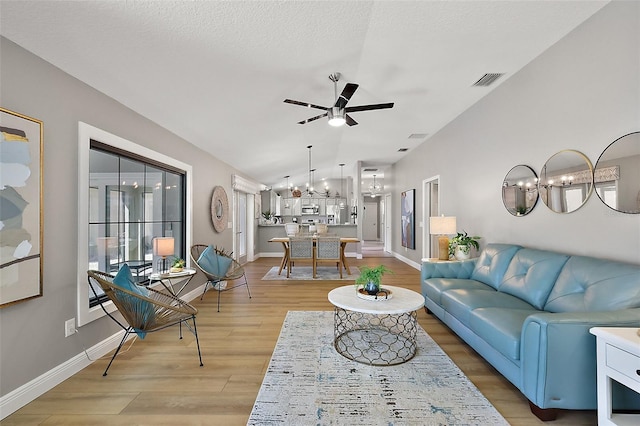 living area featuring a textured ceiling, ceiling fan with notable chandelier, baseboards, vaulted ceiling, and light wood-type flooring