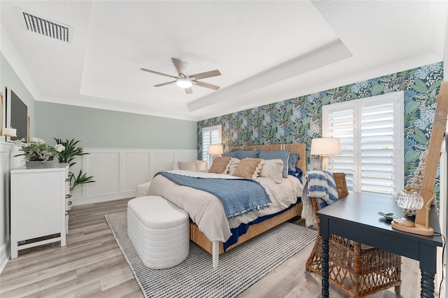 bedroom with wallpapered walls, light wood finished floors, visible vents, a raised ceiling, and wainscoting