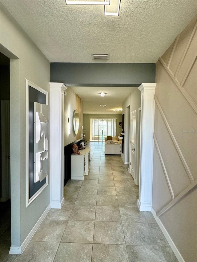 hall with light tile patterned floors, ornate columns, visible vents, a textured ceiling, and baseboards