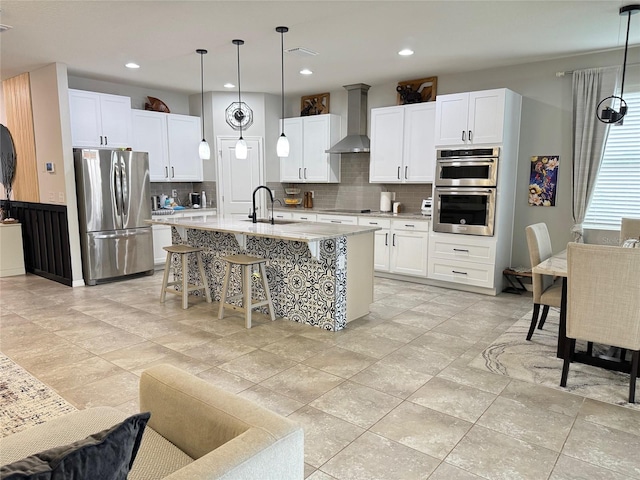 kitchen with stainless steel appliances, hanging light fixtures, white cabinets, wall chimney range hood, and an island with sink