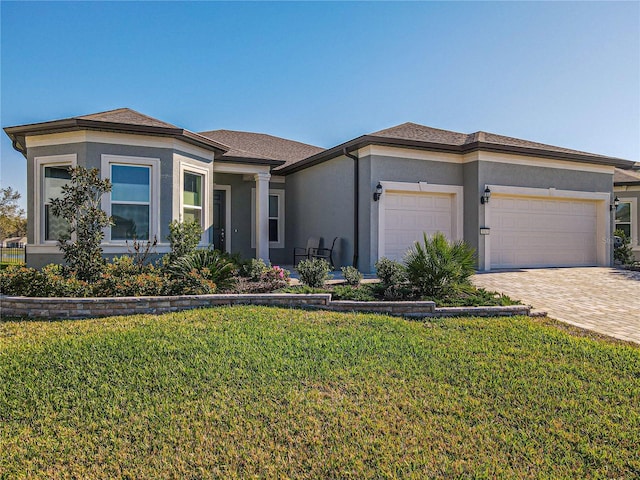 prairie-style house with an attached garage, a shingled roof, decorative driveway, stucco siding, and a front lawn