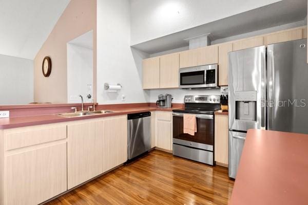 kitchen with light brown cabinets, stainless steel appliances, a peninsula, wood finished floors, and a sink