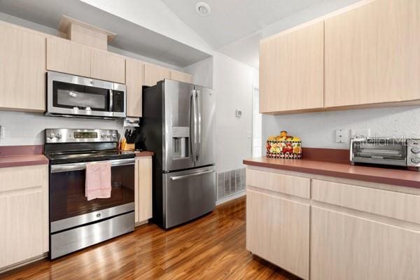 kitchen featuring visible vents, lofted ceiling, appliances with stainless steel finishes, wood finished floors, and light brown cabinets