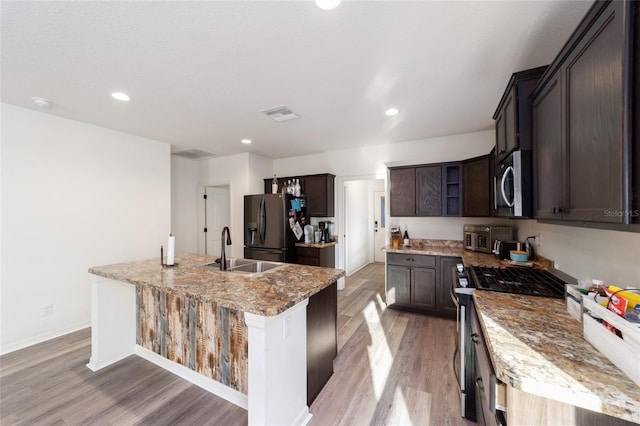 kitchen featuring dark brown cabinetry, a center island with sink, light stone counters, stainless steel appliances, and a sink