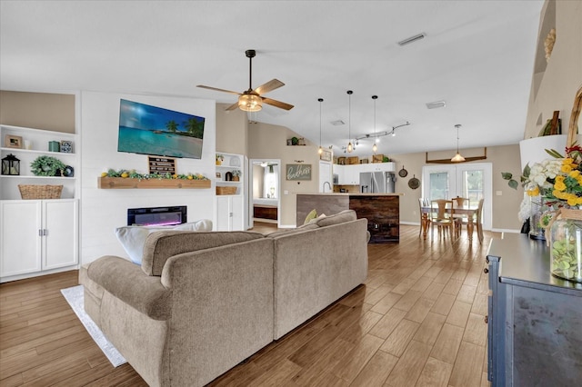 living room featuring a ceiling fan, visible vents, vaulted ceiling, light wood-style floors, and a glass covered fireplace