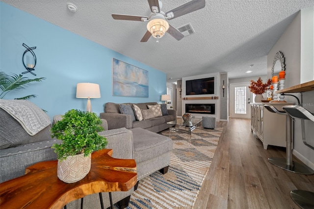 living area featuring a textured ceiling, wood finished floors, visible vents, a ceiling fan, and a glass covered fireplace