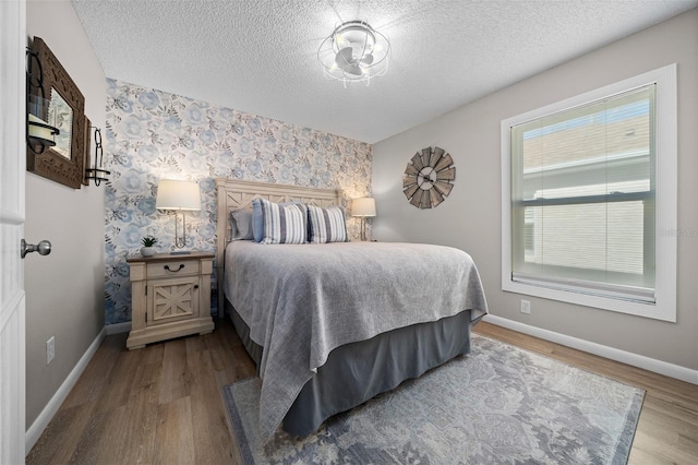 bedroom with a textured ceiling, an accent wall, wood finished floors, baseboards, and wallpapered walls