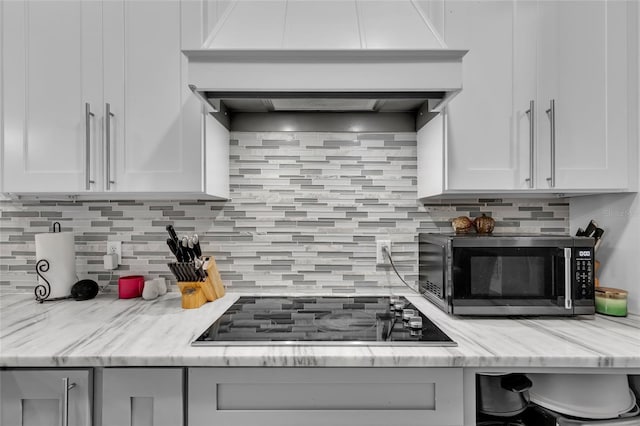 kitchen with tasteful backsplash, custom range hood, stainless steel microwave, black electric stovetop, and white cabinetry