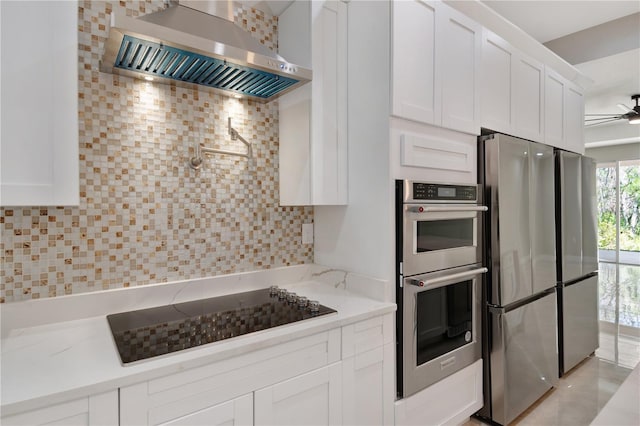 kitchen featuring stainless steel appliances, tasteful backsplash, a ceiling fan, white cabinetry, and wall chimney range hood