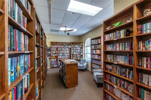 interior space featuring carpet floors, bookshelves, ceiling fan, and a drop ceiling