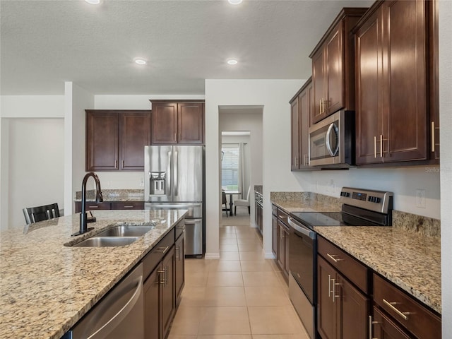 kitchen with light tile patterned floors, dark brown cabinetry, a sink, appliances with stainless steel finishes, and light stone countertops