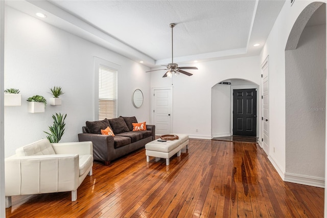 living room with arched walkways, wood-type flooring, a raised ceiling, a ceiling fan, and baseboards