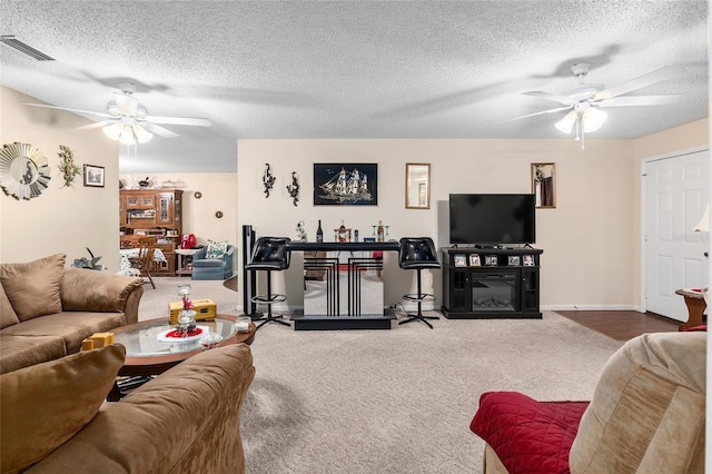 carpeted living area with a textured ceiling, visible vents, a ceiling fan, and a glass covered fireplace