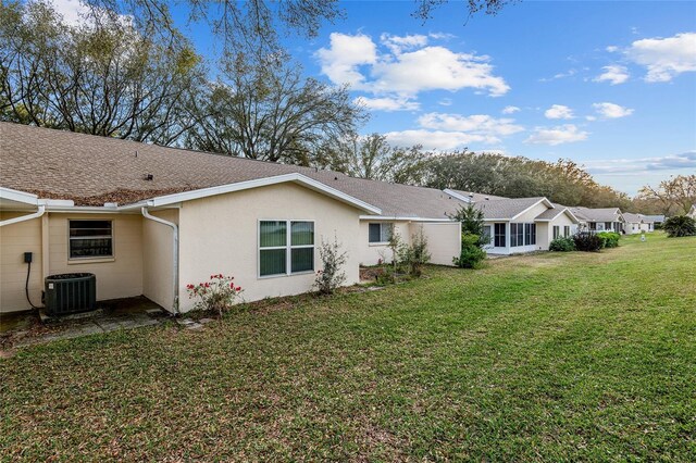 rear view of house with a shingled roof, stucco siding, a yard, and central AC unit