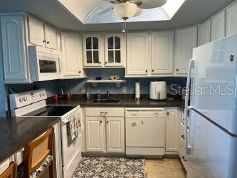 kitchen featuring white appliances, a sink, white cabinets, dark countertops, and glass insert cabinets