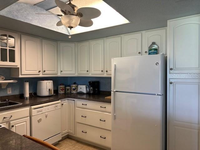 kitchen featuring dark countertops, white cabinets, ceiling fan, a sink, and white appliances