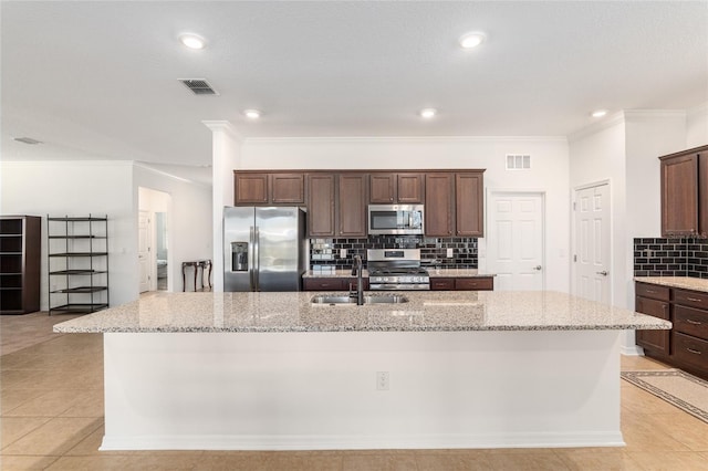 kitchen with light tile patterned floors, stainless steel appliances, a sink, light stone countertops, and an island with sink