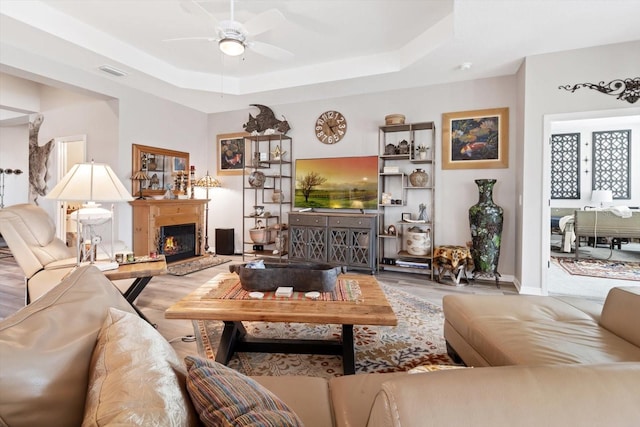 living area featuring visible vents, a raised ceiling, a ceiling fan, wood finished floors, and a lit fireplace