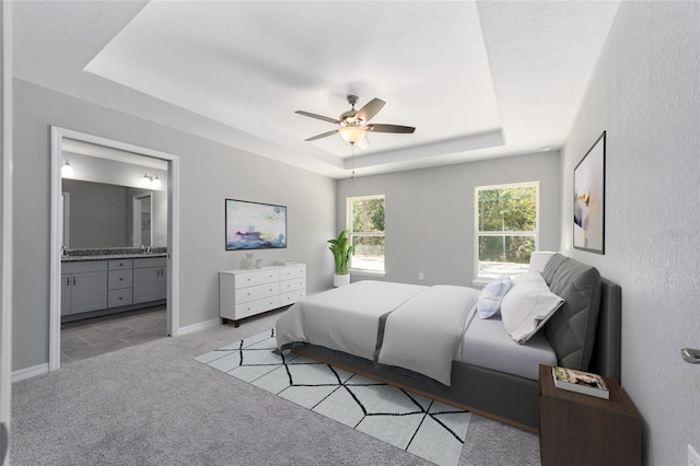 bedroom featuring baseboards, ceiling fan, ensuite bathroom, a raised ceiling, and light colored carpet