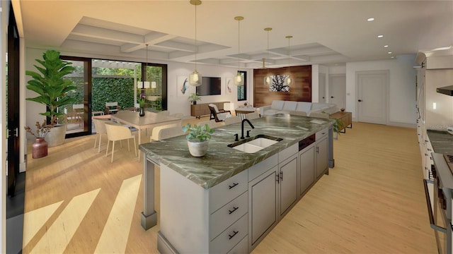 kitchen with a kitchen island with sink, coffered ceiling, a sink, light wood-type flooring, and dark stone countertops