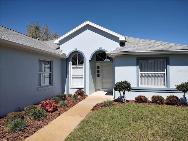 view of exterior entry featuring roof with shingles, a lawn, and stucco siding