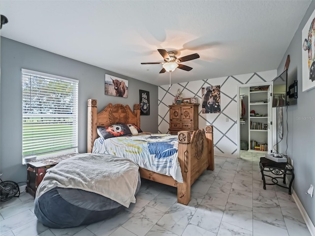 bedroom with a walk in closet, marble finish floor, an accent wall, ceiling fan, and baseboards