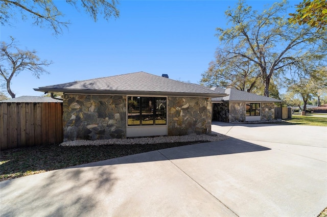 view of front facade featuring stone siding, roof with shingles, fence, and driveway