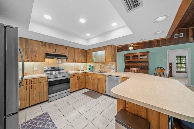 kitchen with under cabinet range hood, a sink, stainless steel appliances, a peninsula, and a raised ceiling